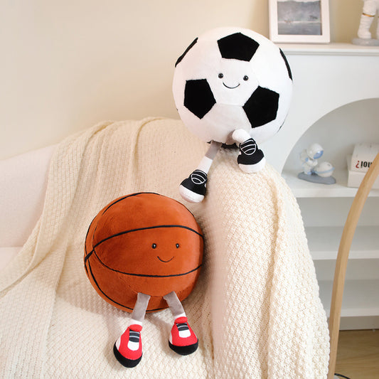Two smiling plush toys shaped like a soccer ball and a basketball are sitting on a cozy white blanket draped over a couch. Both have legs and shoes, giving a playful appearance.