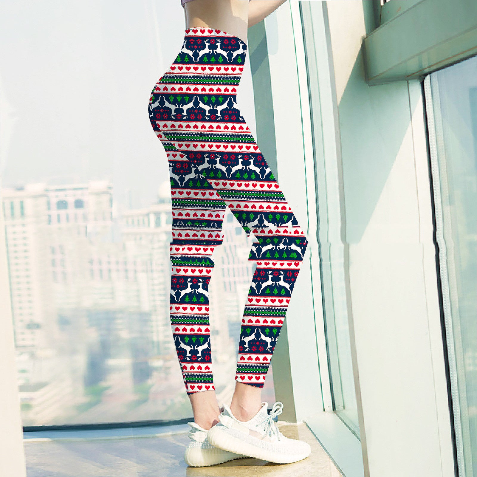 Woman standing by a window wearing festive leggings with a holiday pattern of hearts, trees, and reindeer, paired with white sneakers. Cityscape visible in the background.