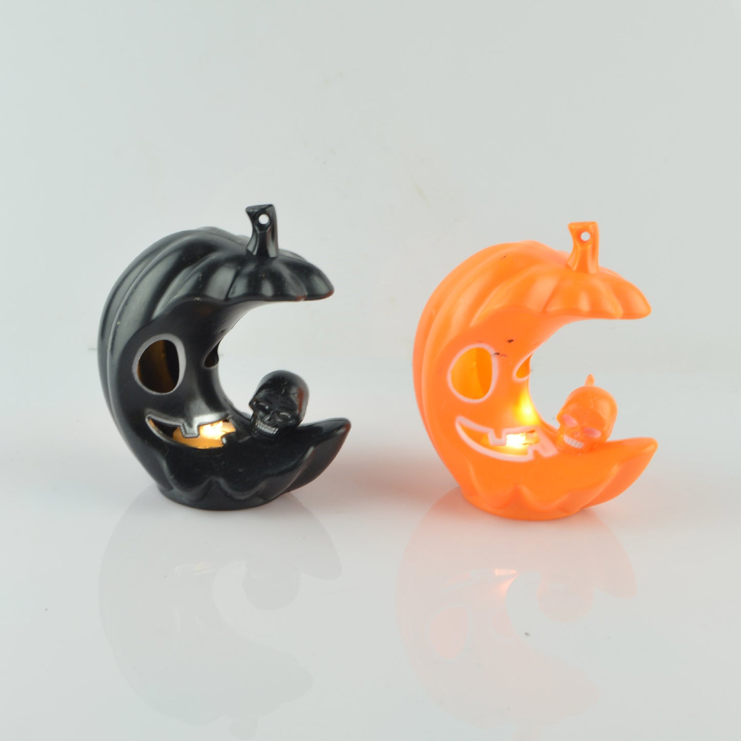 Two crescent-shaped Halloween decorations, one black and one orange, with smiling faces and skulls inside. They emit a soft glow and are set against a white background.
