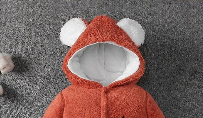 A cozy red infant onesie with a textured finish, featuring a hood with two white fluffy ears, is laid flat on a grey fabric surface. A small portion of a white plush toy is visible on the left.