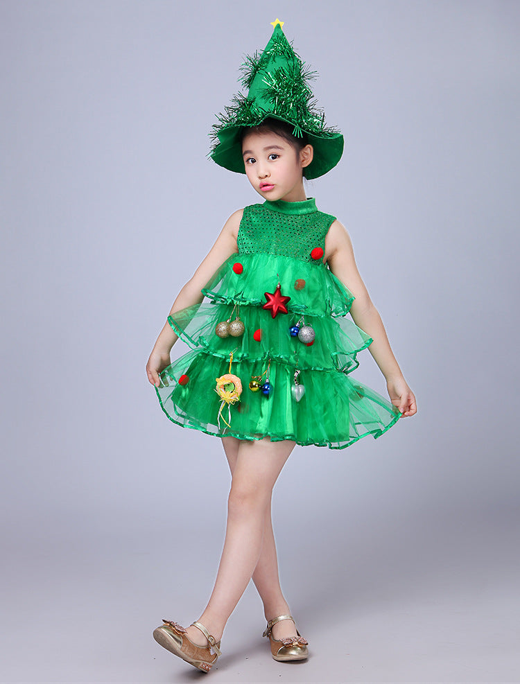 Child wearing a Christmas tree costume with a layered green dress decorated with ornaments. The outfit is topped with a green hat resembling a tree, featuring tinsel and a star.