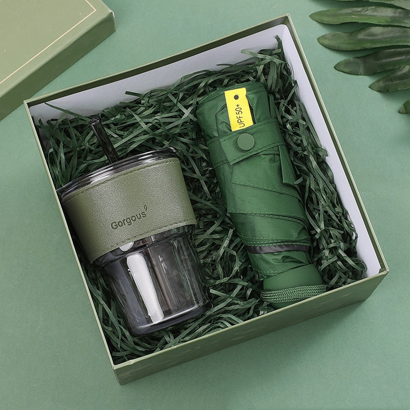 A green gift box containing a clear tumbler with a detachable green sleeve and a folded green umbrella with a "UPF 50+" tag, nestled in shredded green paper.