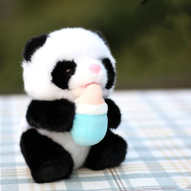 A cute plush panda sits on a checkered cloth, holding a small blue and pink bottle. Its fur is soft and fluffy, with distinctive black patches around its eyes and ears.
