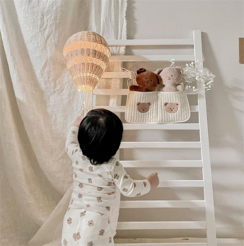 A toddler in bear-patterned pajamas reaches for a woven hot air balloon lamp. Nearby, plush toys sit in a fabric pouch with bear faces on a white wooden rack.