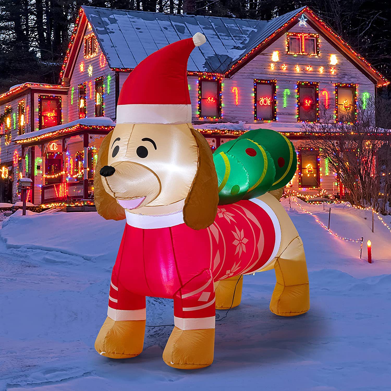 Inflatable dog in a Santa hat and festive attire stands in a snowy yard, with a large candy cane on its back. A brightly lit, decorated house is visible in the background.
