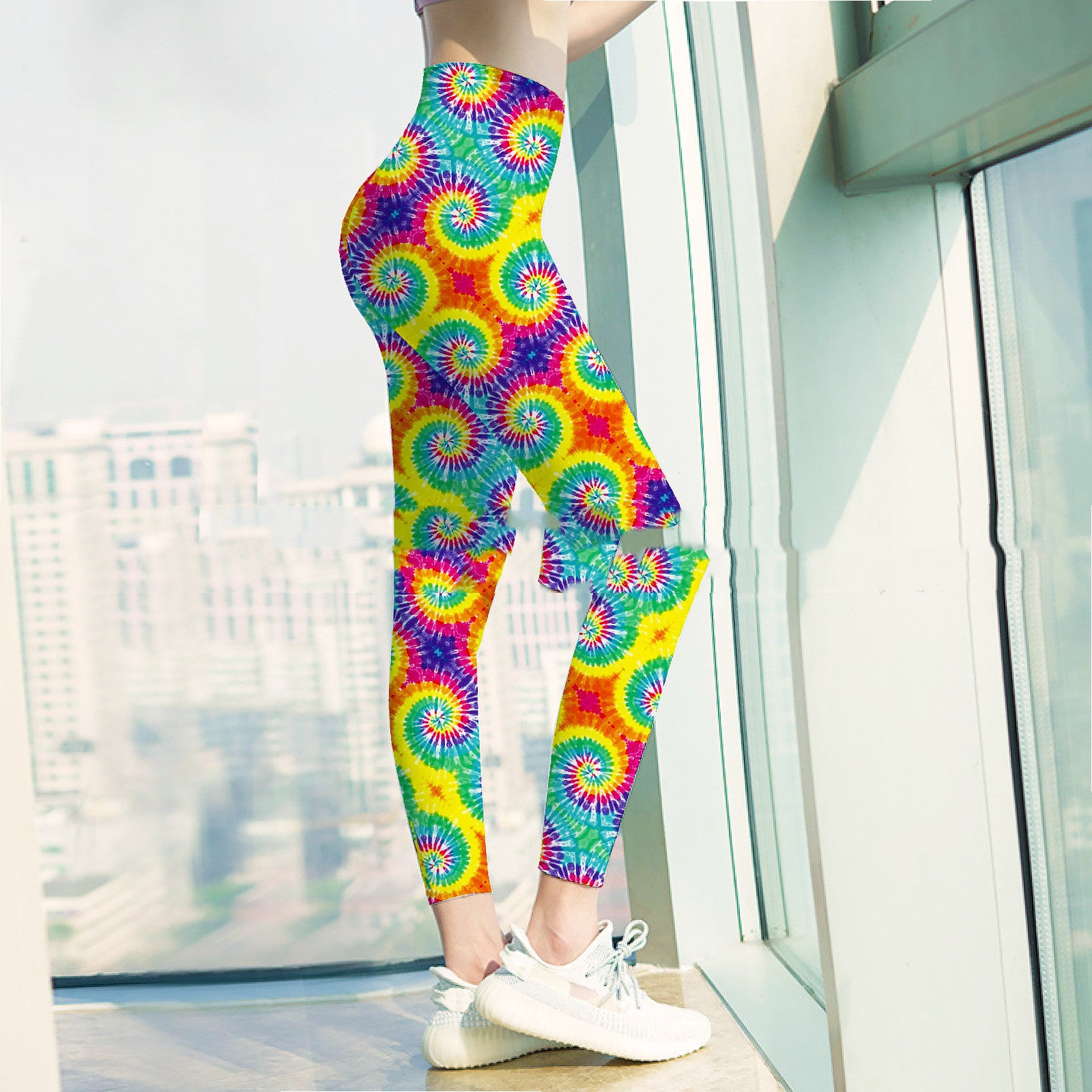 Person in vibrant tie-dye leggings and white sneakers stands by a window, revealing an urban skyline in the background.