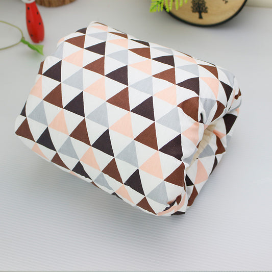 A folded quilt with a pattern of pastel pink, grey, and brown triangles lies on a light surface. Decor includes a small red object and a round wooden item with tree designs.