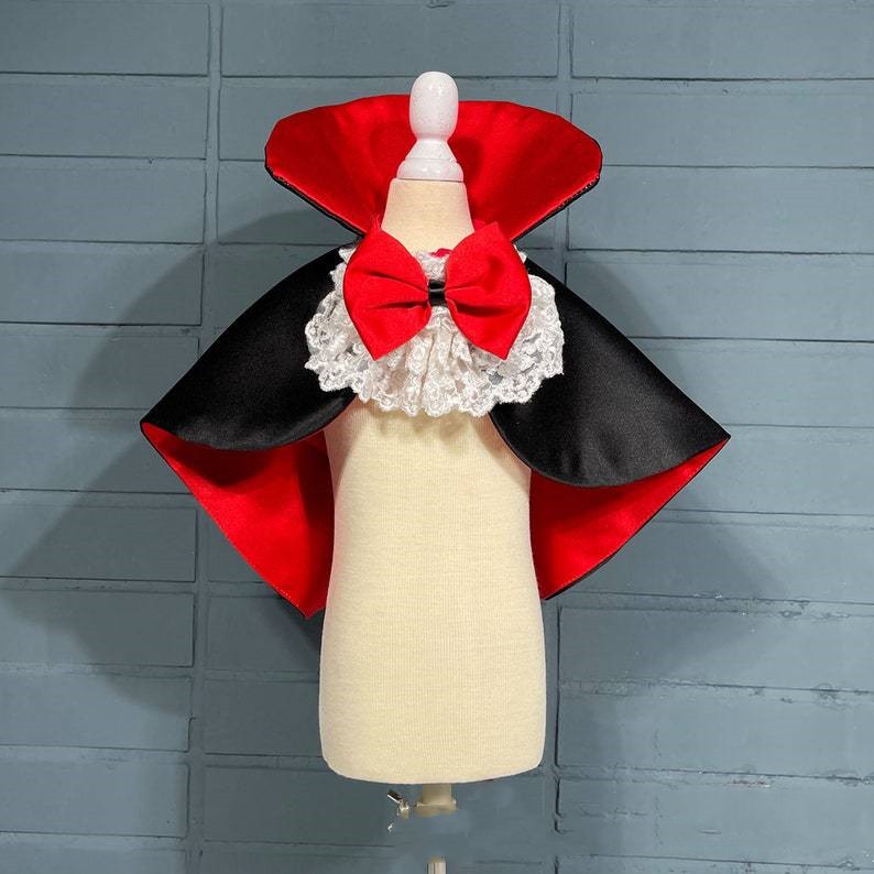 Dog mannequin displays a vampire costume with a black and red cape, frilly white lace collar, and a large red bow, set against a gray brick wall.