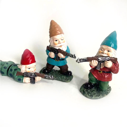 Three garden gnomes, each holding a rifle. One is lying prone in green, while the others stand in blue and red outfits. They have cheerful expressions and point forward.
