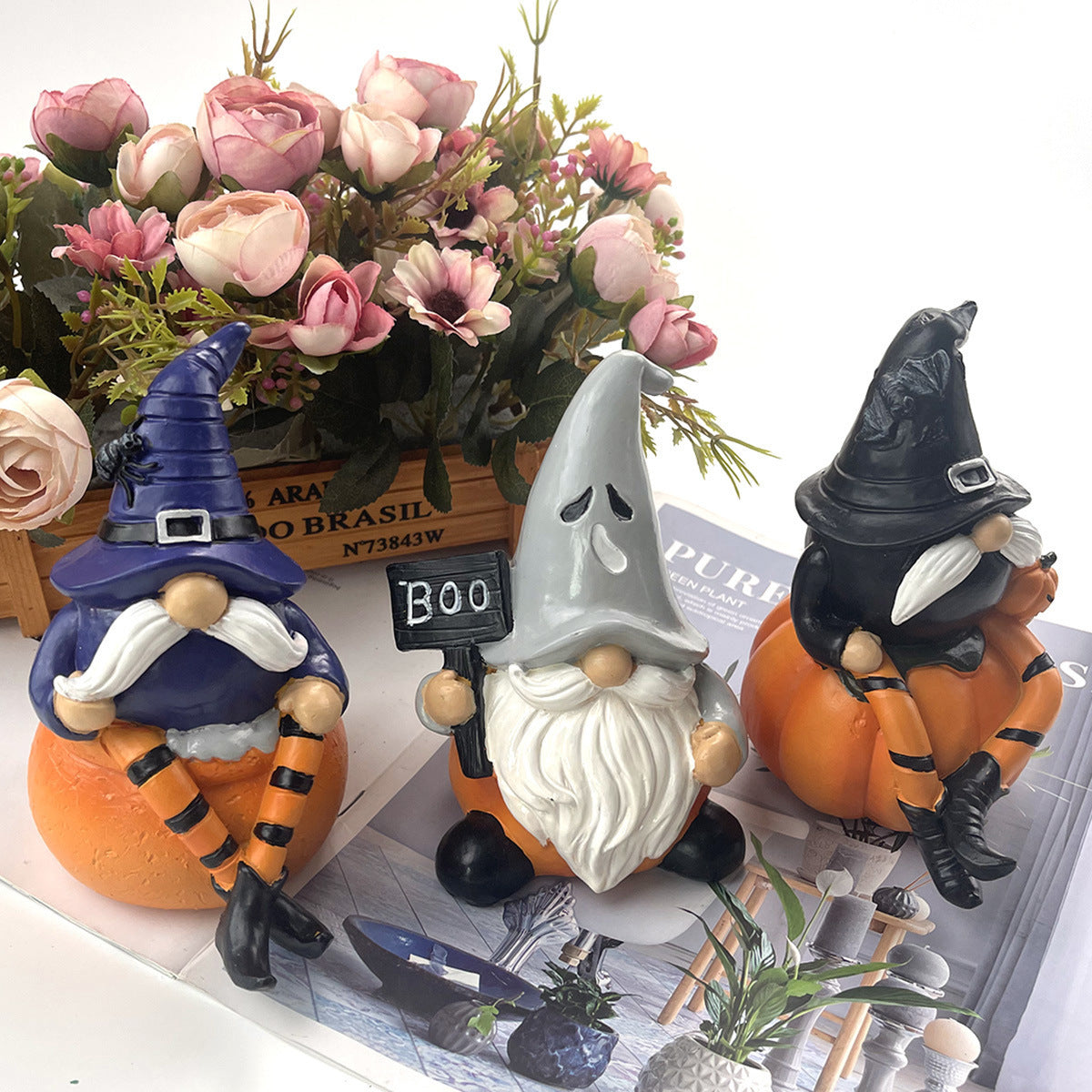 Three whimsical gnome figurines in Halloween costumes sit on pumpkins. One holds a "Boo" sign. Pink flowers in a wooden box are in the background.
