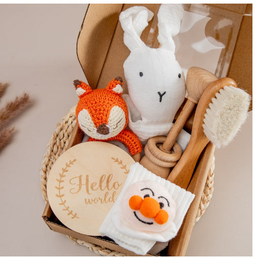 Gift box with a crochet fox, white bunny toy, wooden baby brush, rattle, and cloth with pom-pom smiley. A round sign reads "Hello world" in elegant script.
