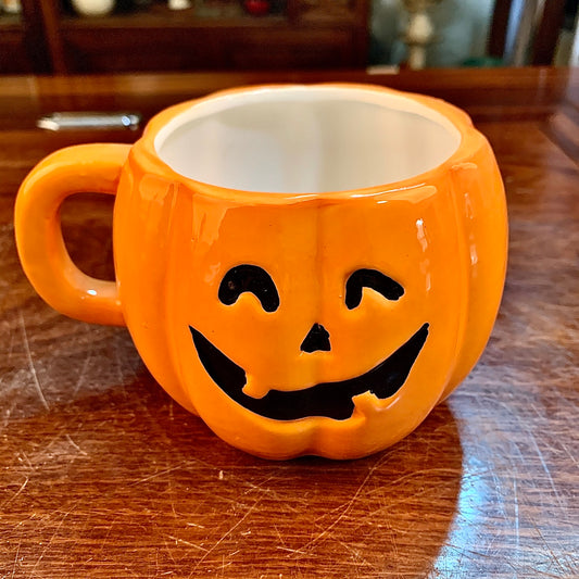 Orange pumpkin-shaped mug with a carved, smiling jack-o'-lantern face, placed on a wooden surface.