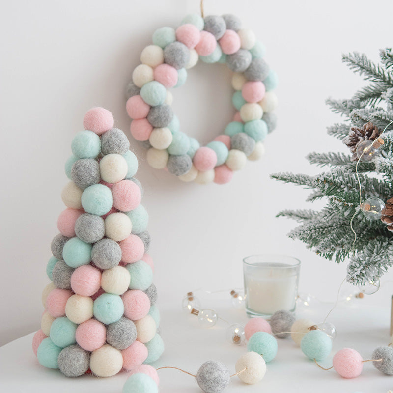 Pastel-colored felt ball wreath and cone tree on white surface, with a frosted mini Christmas tree, glowing string lights, and a candle create a cozy festive scene.