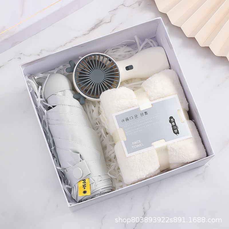 A gift box containing a white towel, a portable fan, and a folded umbrella, all neatly arranged on shredded paper. The setting features a marble-like background.