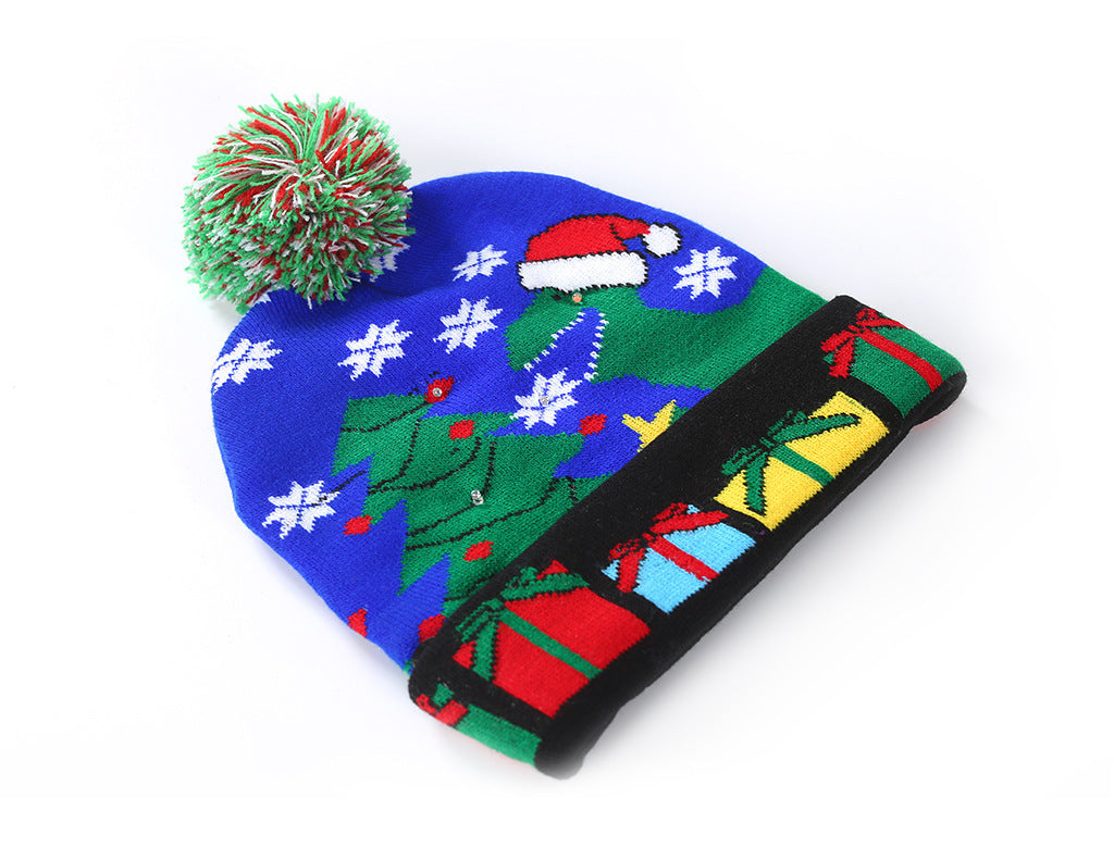Festive Christmas beanie featuring a pom-pom, decorated with a Santa hat, snowflakes, and a tree. The black brim displays colorful wrapped gift illustrations.