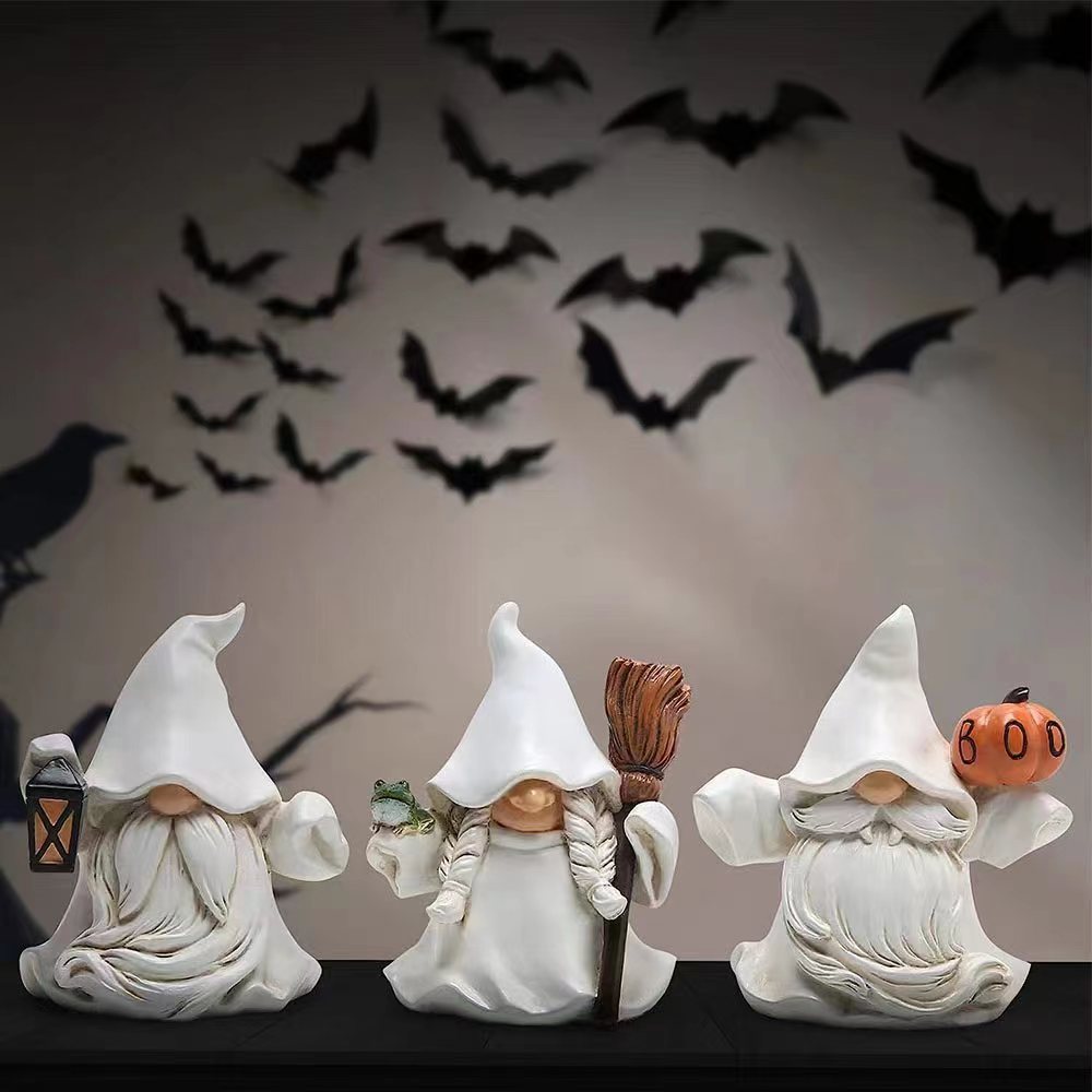 Three gnome figurines in white robes with pointed hats sit on a shelf. Each holds a Halloween-themed object: a lantern, a broom with a frog, and a pumpkin labeled "BOO." Black bats decorate the wall behind.