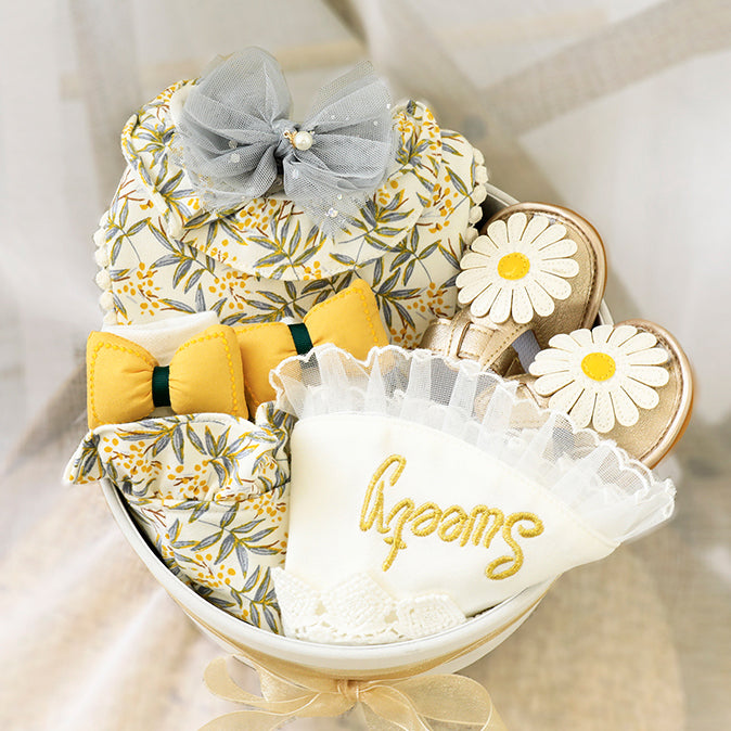 A decorative gift set in a bowl, featuring floral-print fabric items with a gray bow, gold flat shoes with daisy embellishments, and a cloth with “Sweet” embroidered in gold.