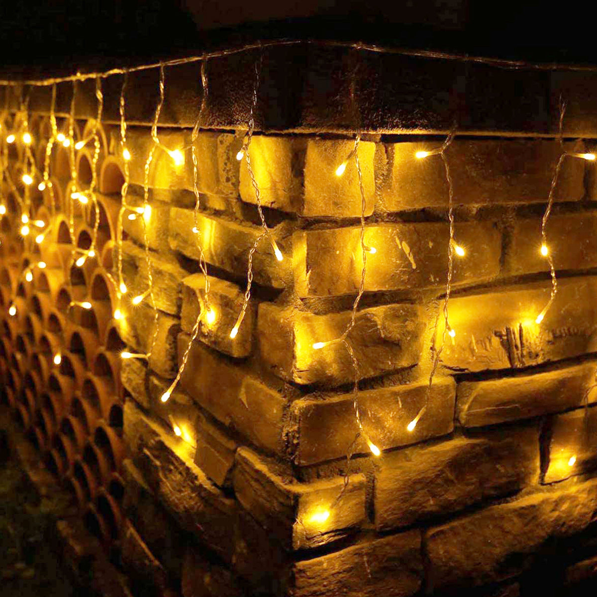 Warm yellow string lights hanging on a textured brick wall, casting a cozy glow. The lights drape down the corner, enhancing the rustic ambiance in the dark.