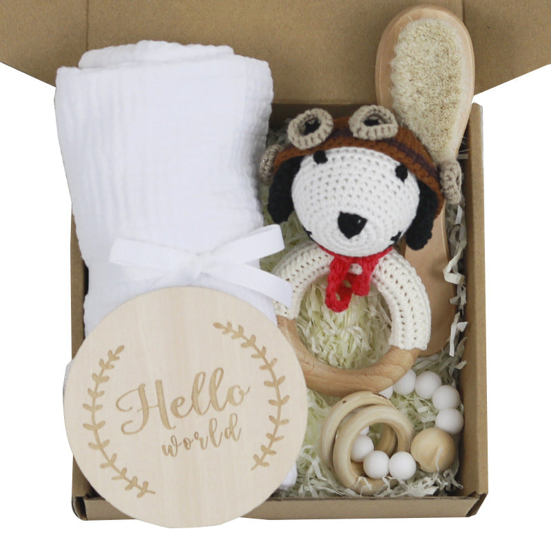 Gift box with a white cloth, crochet dog rattle wearing goggles, wooden massage brush, teething rings, and a "Hello world" plaque, all surrounded by shredded paper.