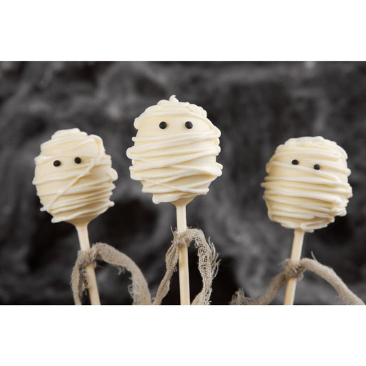 Three mummy cake pops wrapped in white icing with black eyes are arranged in front of a dark, blurred background. Each has frayed brown ribbon tied around the stick.
