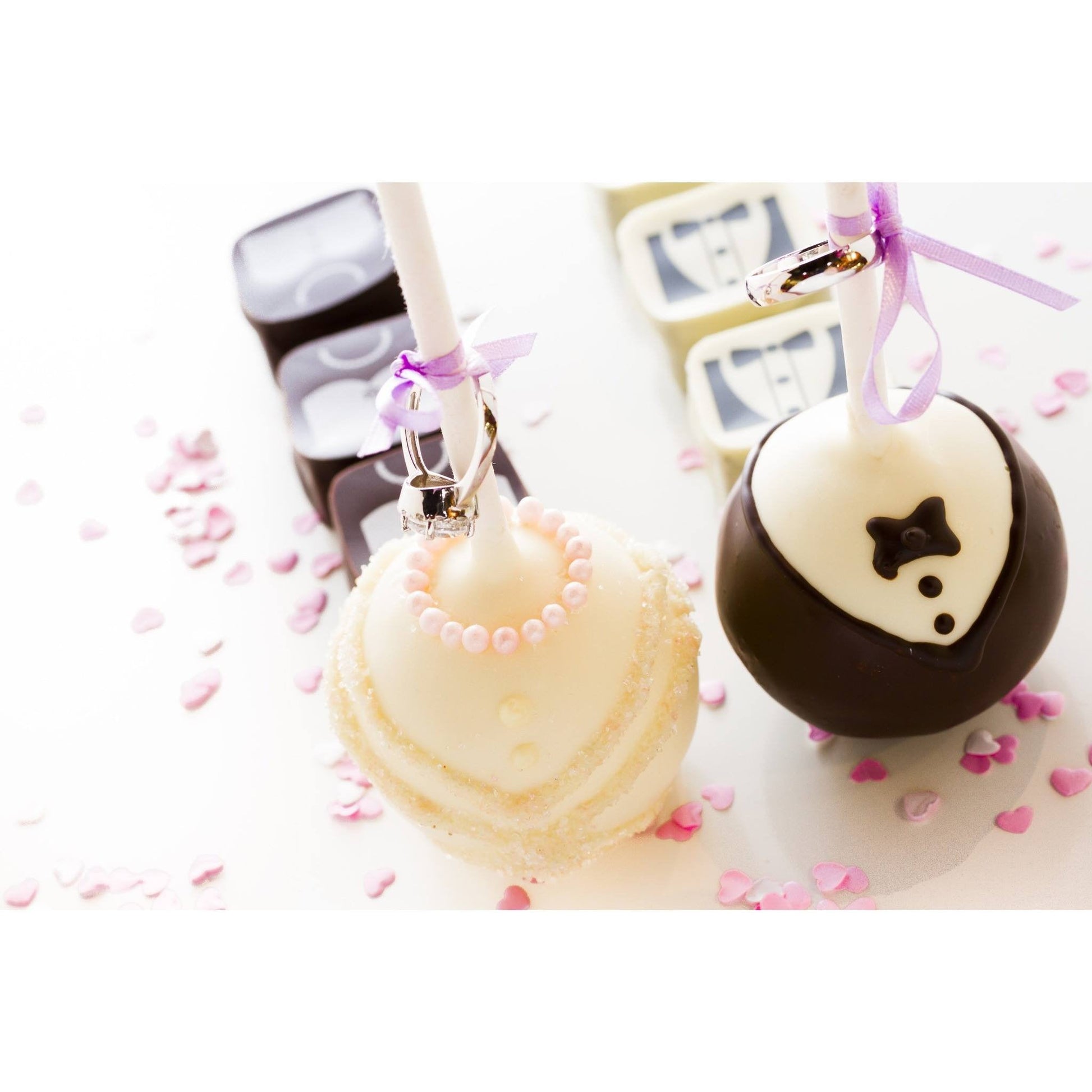 Two cake pops designed as wedding attire, one in white with pink pearls and the other in black and white tuxedo style, adorned with pink ribbons and rings, surrounded by pink heart confetti.