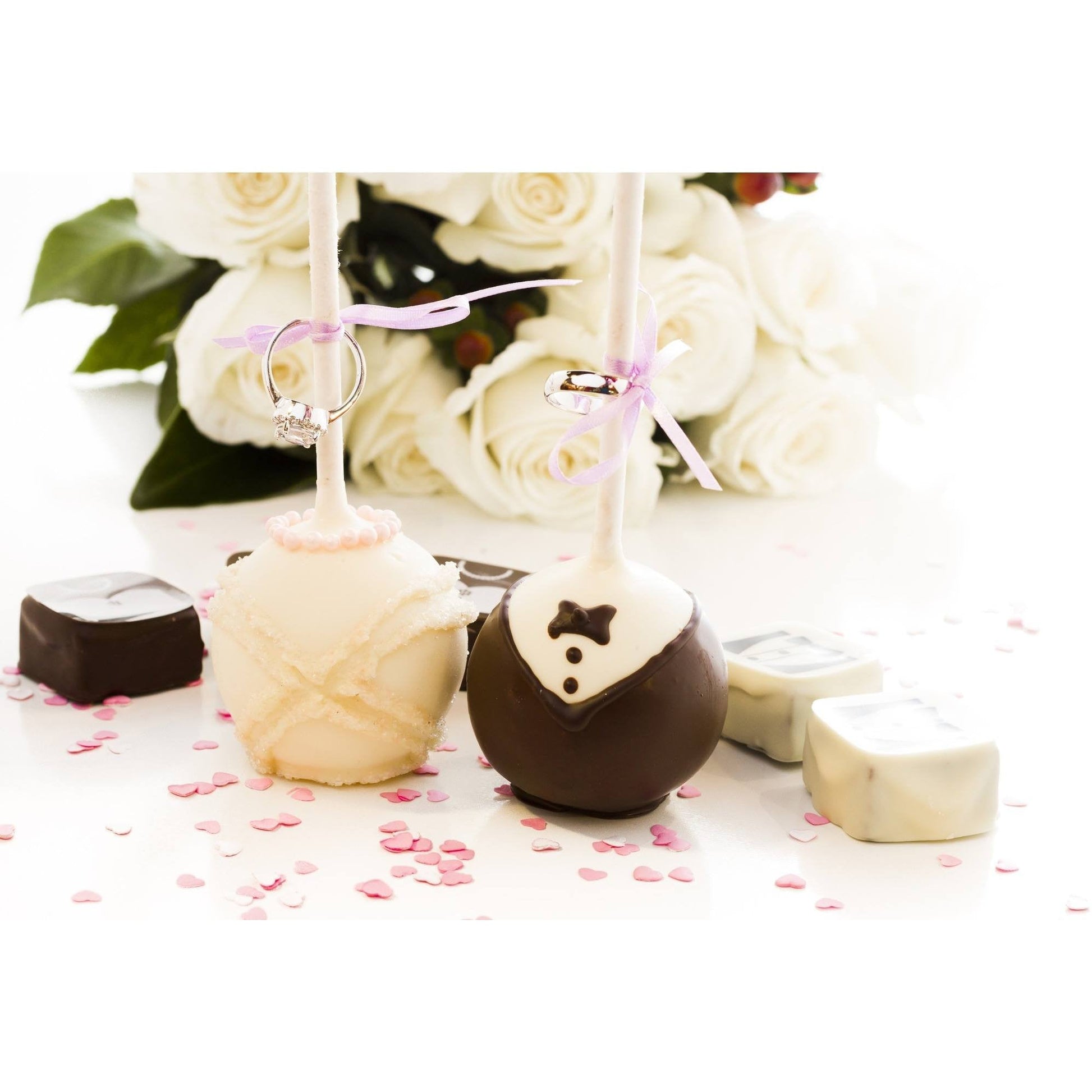 Wedding-themed cake pops designed as a bride in white and a groom in chocolate, adorned with rings, surrounded by rose petals and chocolates, with roses in the background.