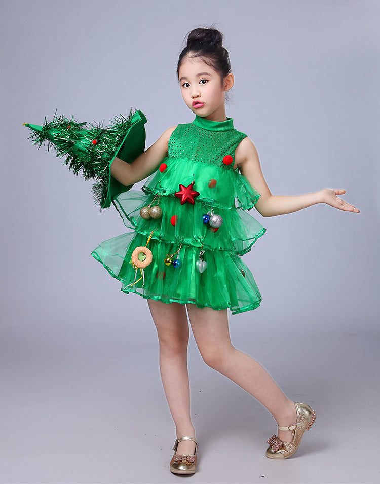 Young girl in festive green dress decorated with ornaments, holding a matching tree-shaped hat. She poses playfully, wearing gold shoes against a plain background.