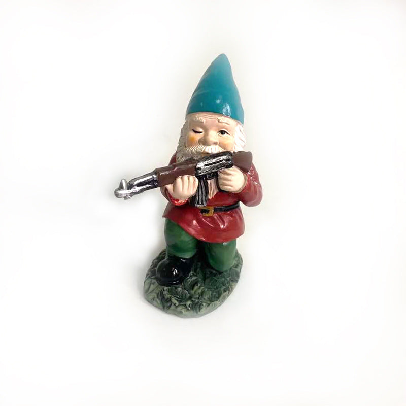 A garden gnome with a blue hat and red coat holds a small toy gun, standing on a grassy base. The figure has a playful, mischievous expression.