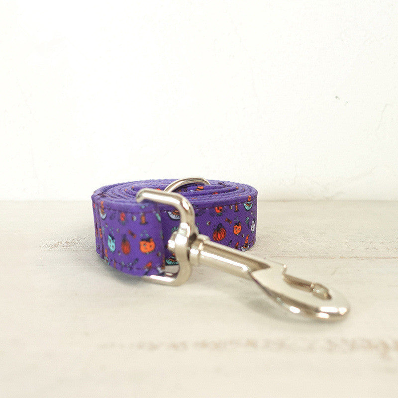 Purple dog leash with Halloween-themed patterns of pumpkins and ghosts, featuring a shiny metal clasp, placed on a light wooden surface.