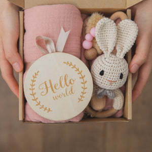 Hands holding a gift box containing a pink blanket, a wooden plaque with "Hello world" text, and a crocheted bunny toy with wooden beads.