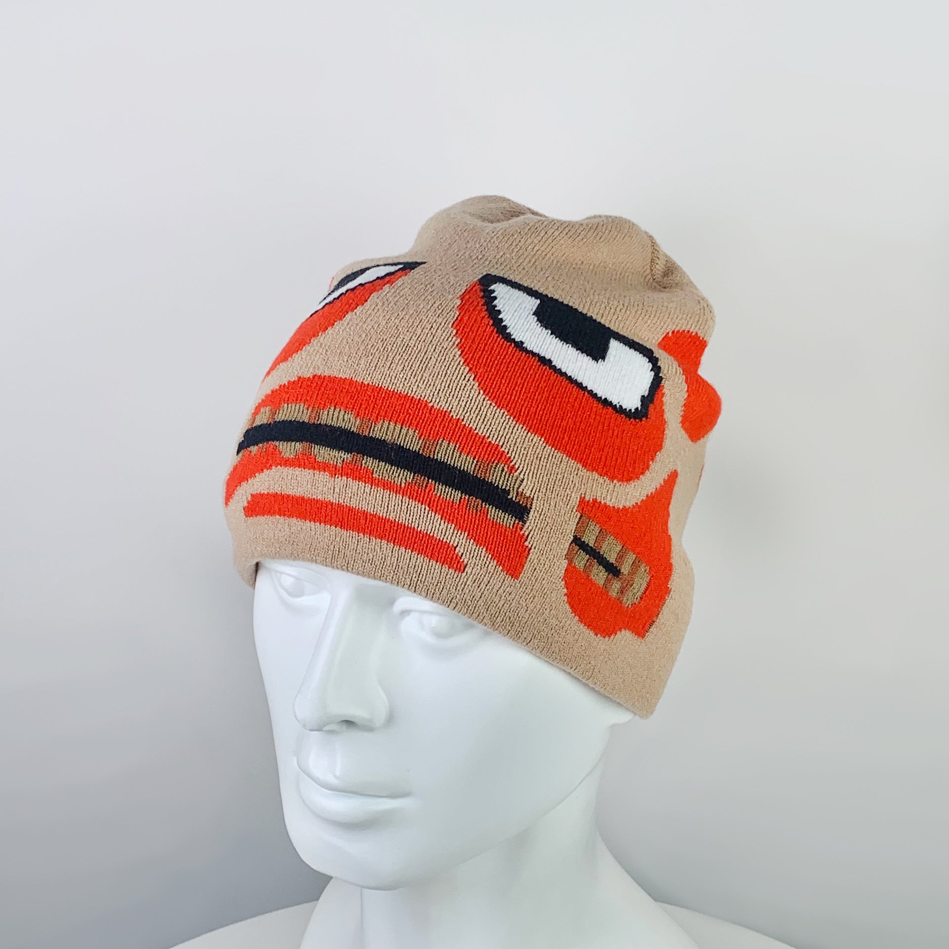 Beige beanie with playful cartoon face design in orange and black, featuring large eyes and expressive mouth. Displayed on a white mannequin head against a plain background.