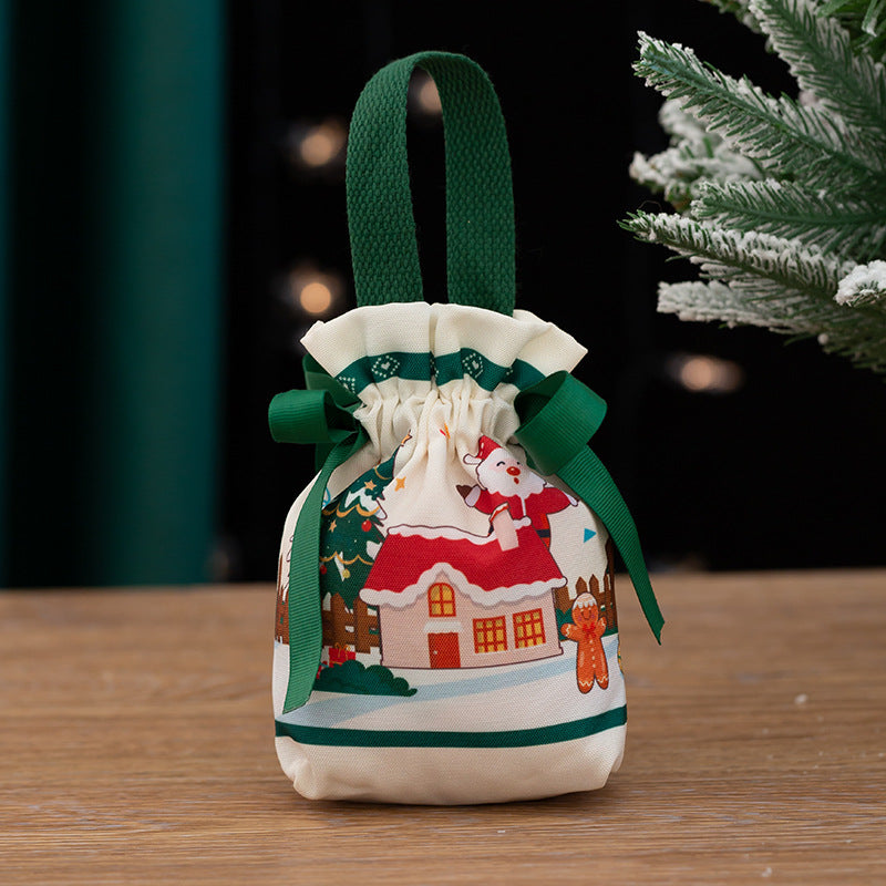 Festive fabric gift bag with green handles, featuring a holiday scene of Santa, a gingerbread man, and a snowy house, set against a wooden table and a blurred festive backdrop.