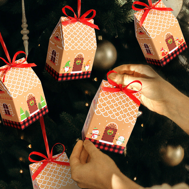 Hands decorating a Christmas tree with small, house-shaped gift boxes. The boxes have festive designs, red ribbons, and are hung among dark green branches and soft lights.
