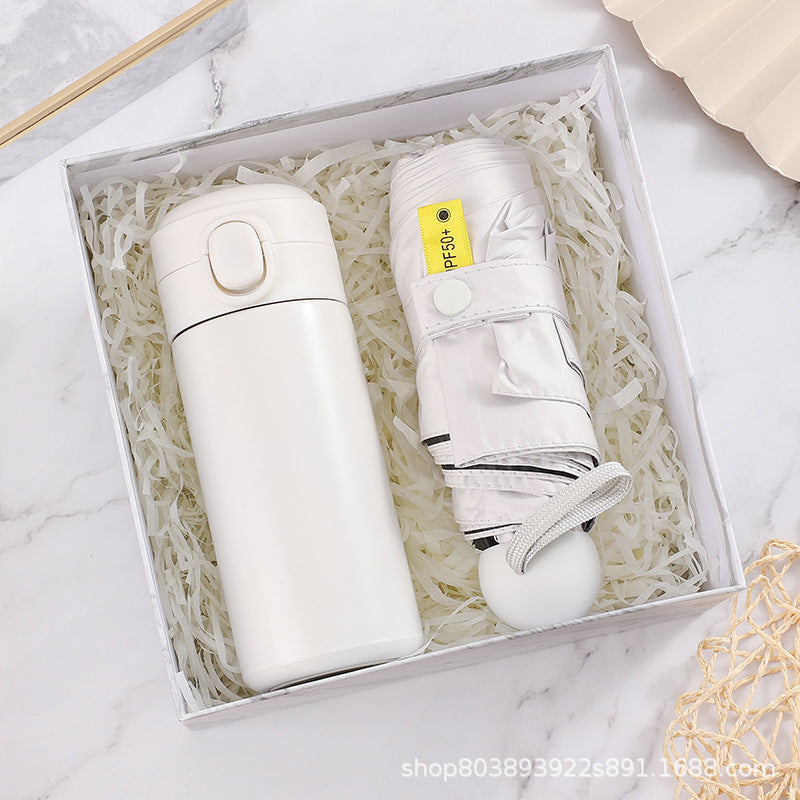 White thermos and folded umbrella with a "UPF 50+" label, placed on shredded paper in a white gift box on a marble surface.