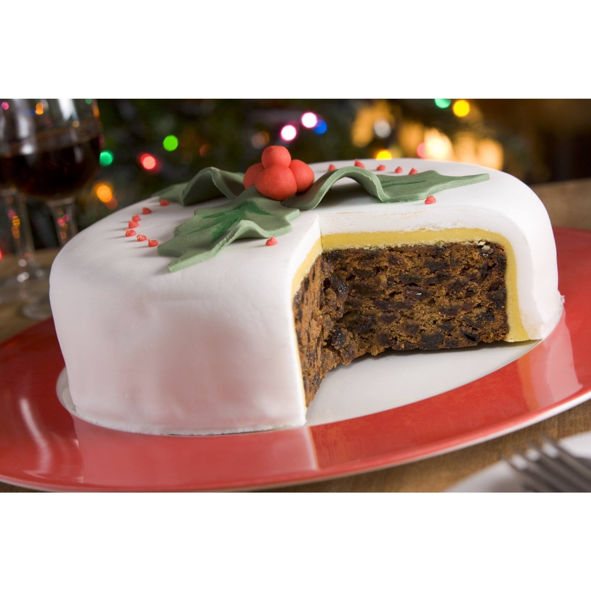 Festive fruitcake with white icing and holly decorations on a red plate, a slice removed to reveal rich, dark interior. Blurred colorful lights in the background.