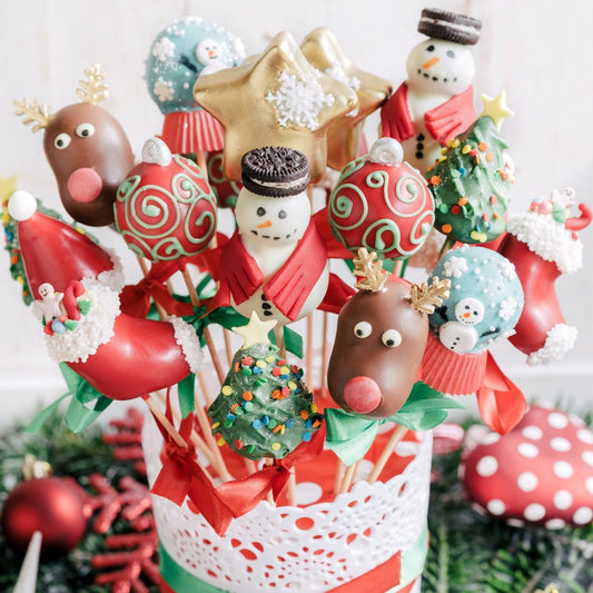 Festive cake pops featuring snowmen, reindeer, Christmas trees, Santas, and ornaments in vibrant holiday colors, arranged in a decorative white and red container with green accents.