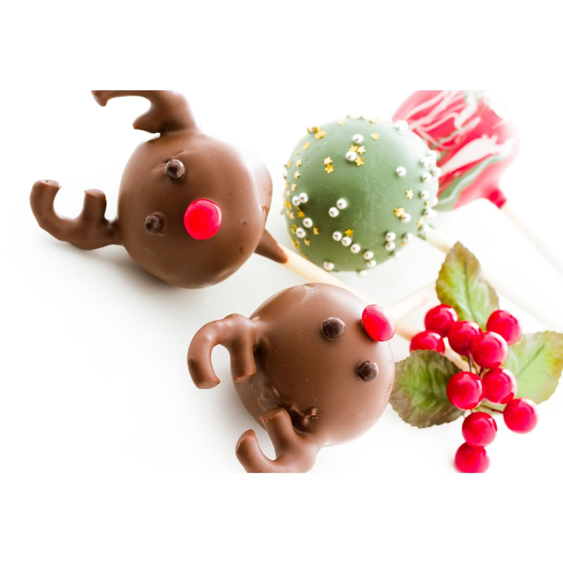 Festive cake pops decorated as reindeer with chocolate antlers and red noses, a green pop with silver and gold beads, and a red swirled pop, alongside red berries and leaves.