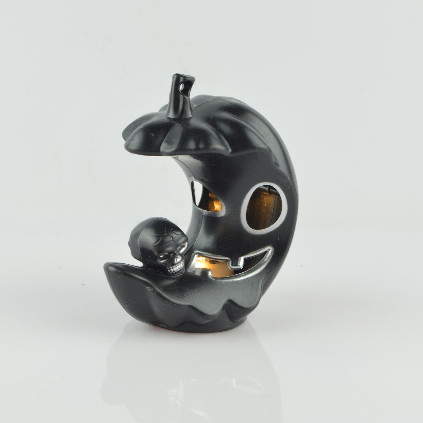 A black, crescent-shaped ceramic pumpkin with a carved face, featuring a small skull nestled at the base, set against a plain white background.