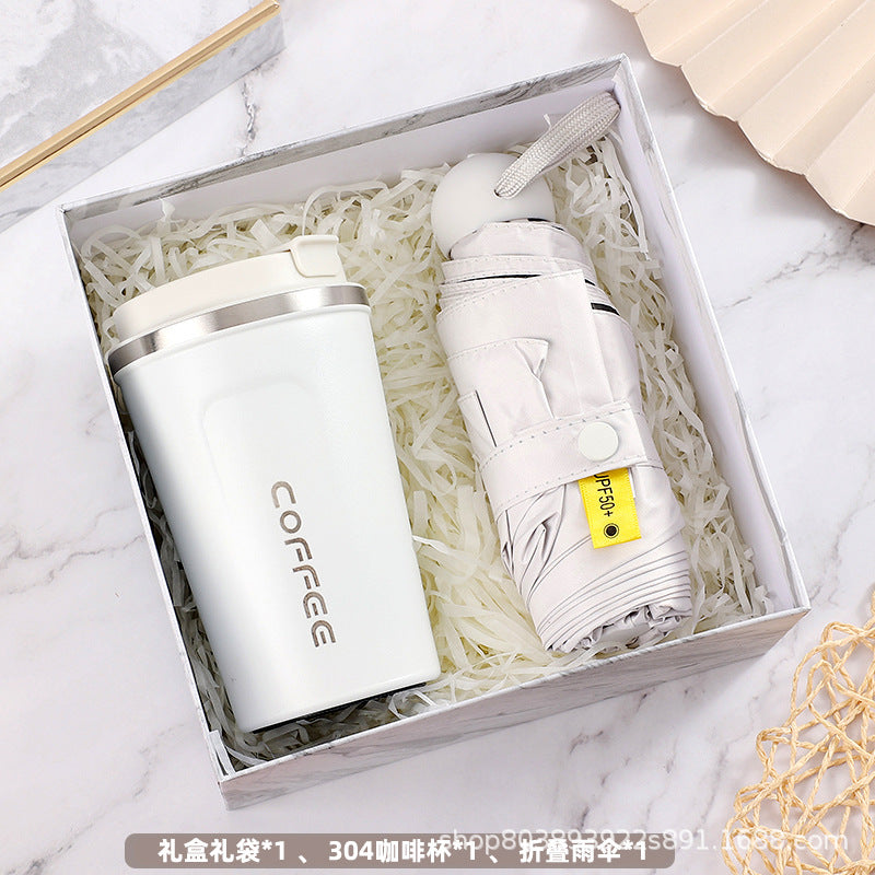 Gift box containing a white insulated coffee tumbler and a compact folded umbrella, both nestled in shredded white paper on a marble surface.