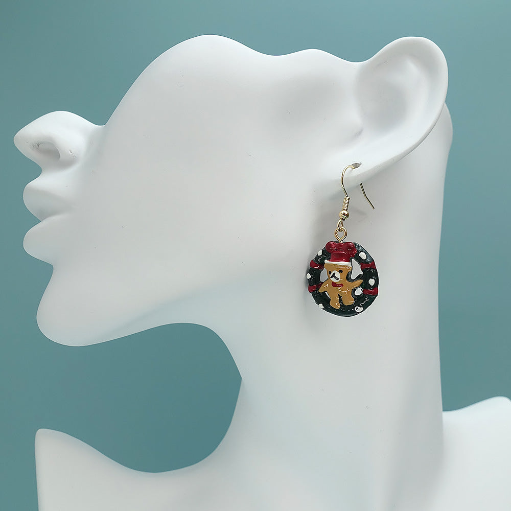 A mannequin head displays a festive earring featuring a gingerbread man with a red hat, surrounded by a green wreath with black and white accents, on a light blue background.