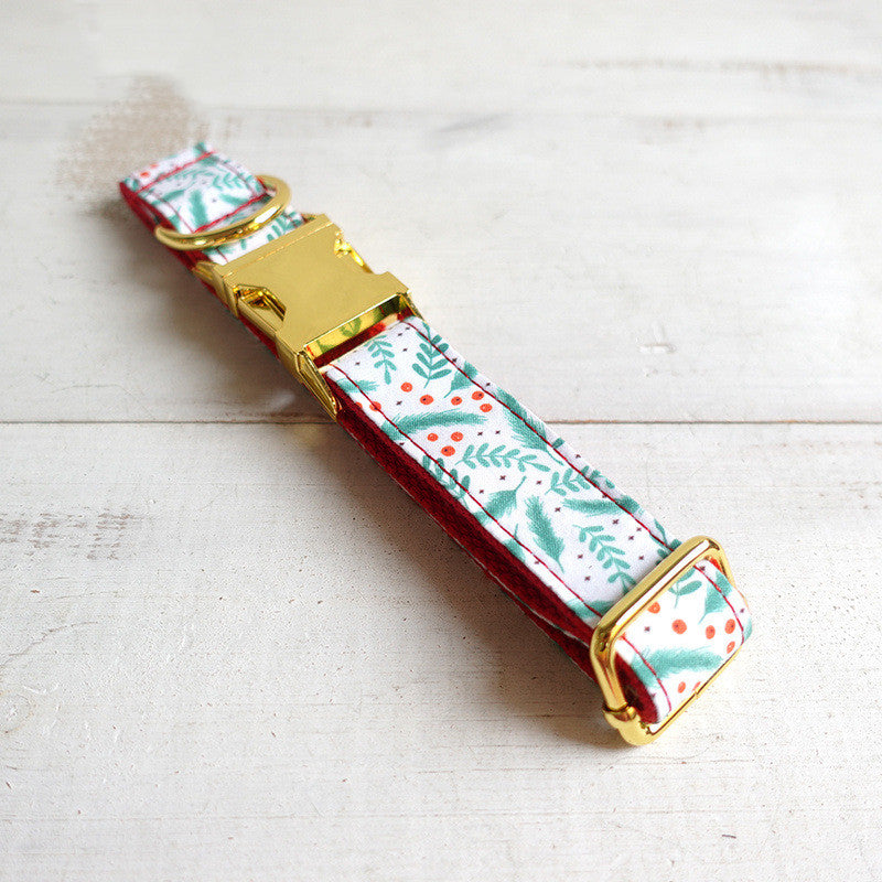 Festive dog collar with a gold buckle, featuring a white background with green foliage and red berries pattern, laid on a light wooden surface.