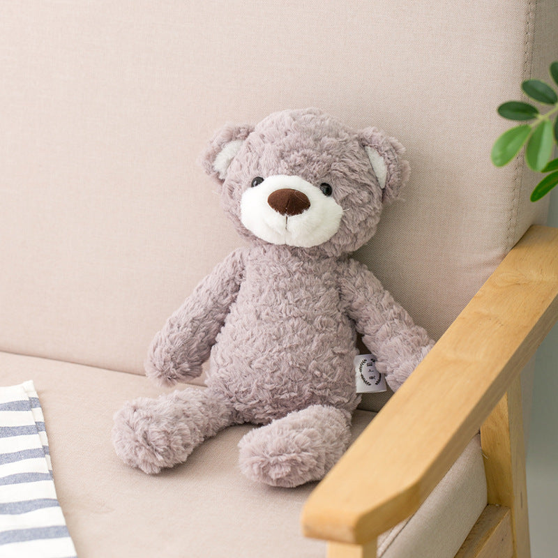 A plush gray teddy bear with a white snout and brown nose sits on a beige sofa next to a striped fabric and a wooden armrest. A small green plant is visible nearby.