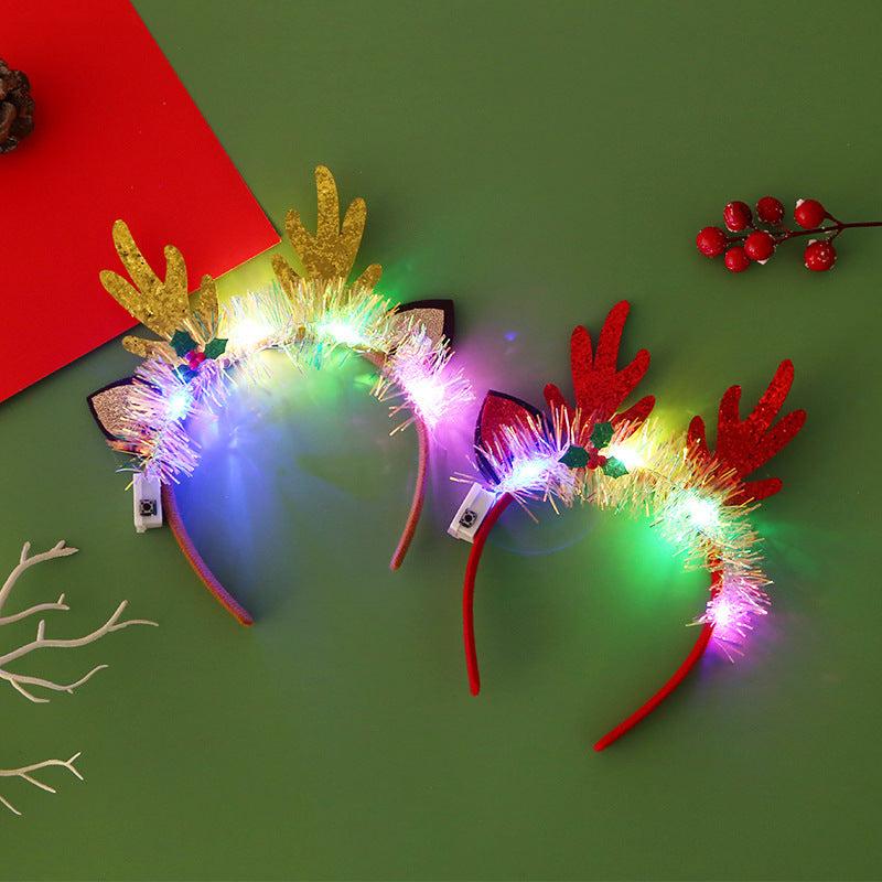 Two festive reindeer antler headbands, one gold and one red, adorned with colorful lights and tinsel, lie on a green surface with red berries and a red paper beside them.