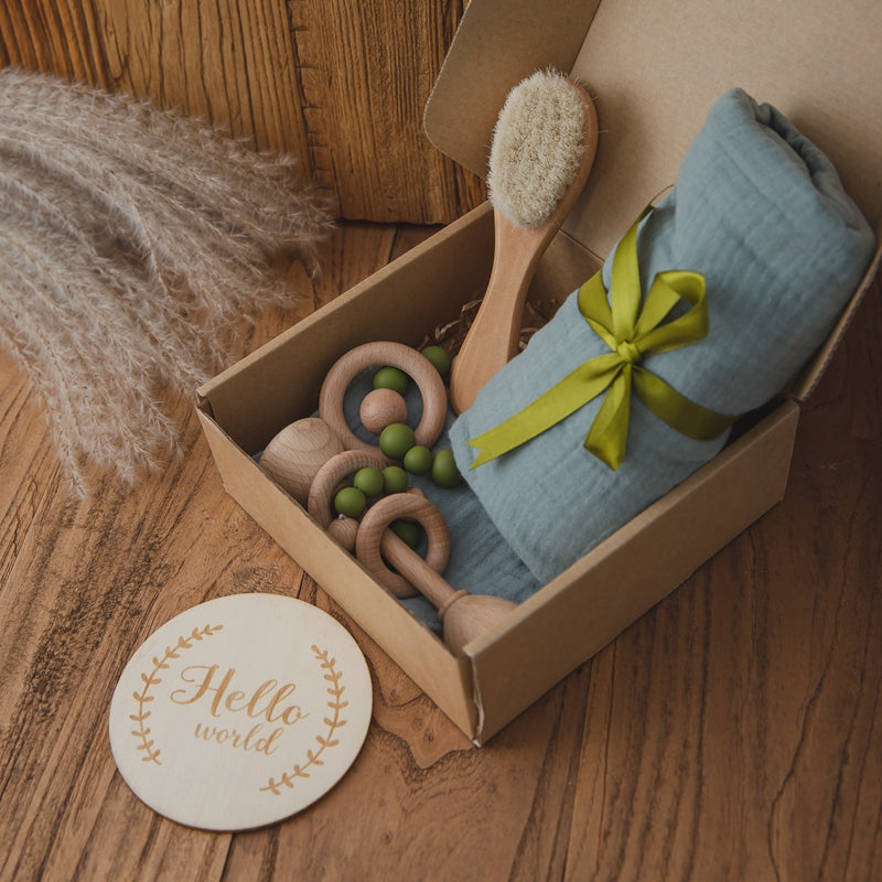 A cardboard box contains a blue fabric roll tied with a green ribbon, wooden rattles, and a brush. Nearby, a wooden plaque reads "Hello world," on a wooden surface with pampas grass.