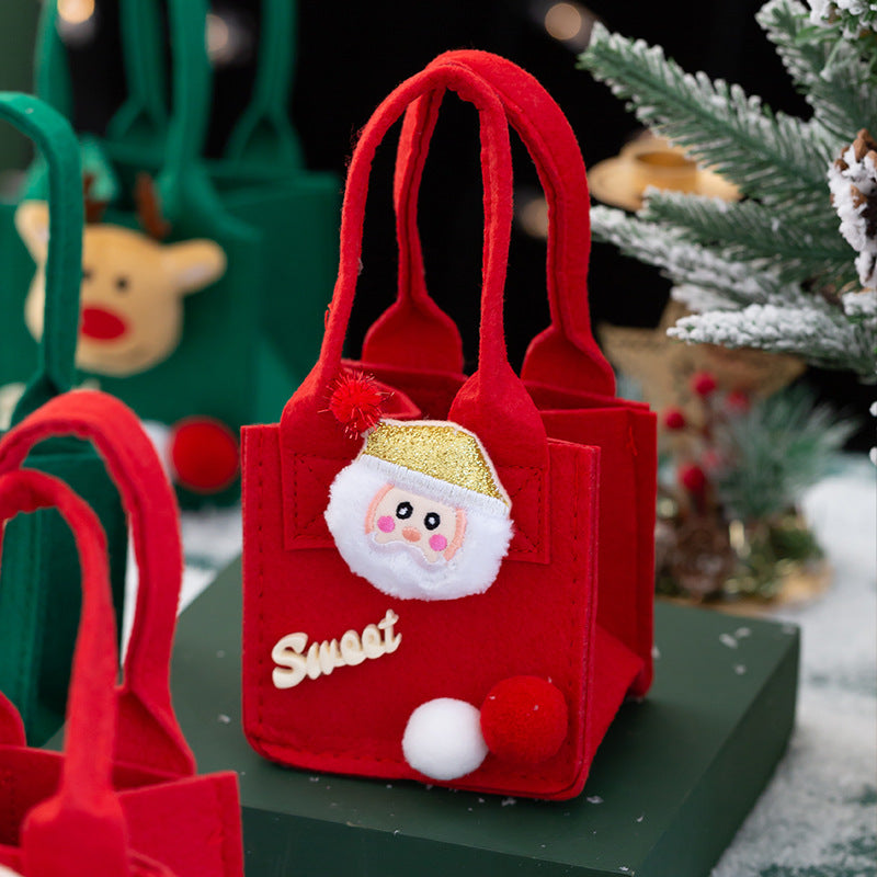 Red felt gift bag with a Santa face and pom-poms, featuring the word "Sweet." A green bag with a reindeer design is in the background, with snow-covered branches nearby.