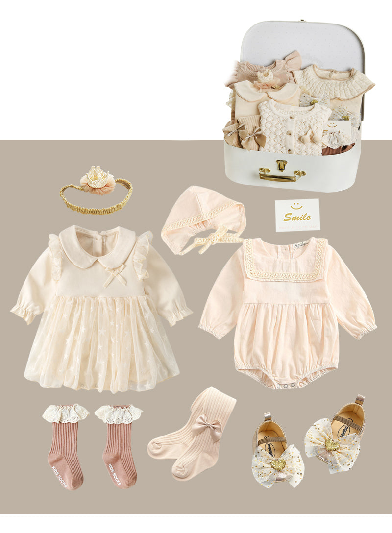 Set of beige baby clothes including a dress, romper, socks, bonnet, headband, and shoes with gold accents, neatly arranged with a suitcase and "Smile" card.