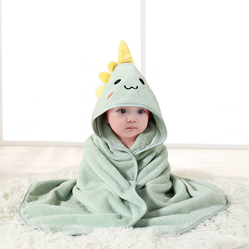 A baby wrapped in a soft green blanket with a dinosaur hood, complete with yellow spikes and a cute face design, sits on a fluffy white rug in a well-lit room.