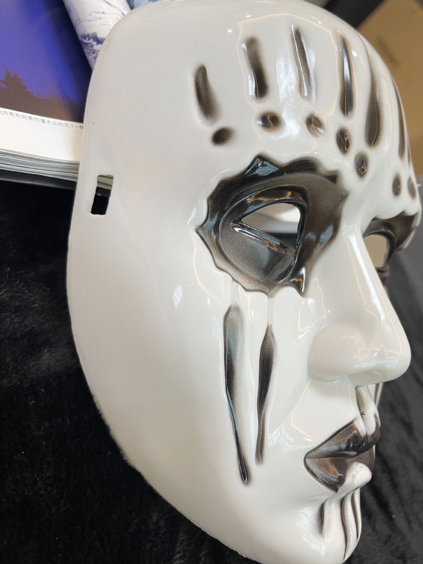 White ceramic mask with black abstract markings around the eyes, forehead, and mouth, lying on a black surface. Partial view of an open book with a blue cover is visible.