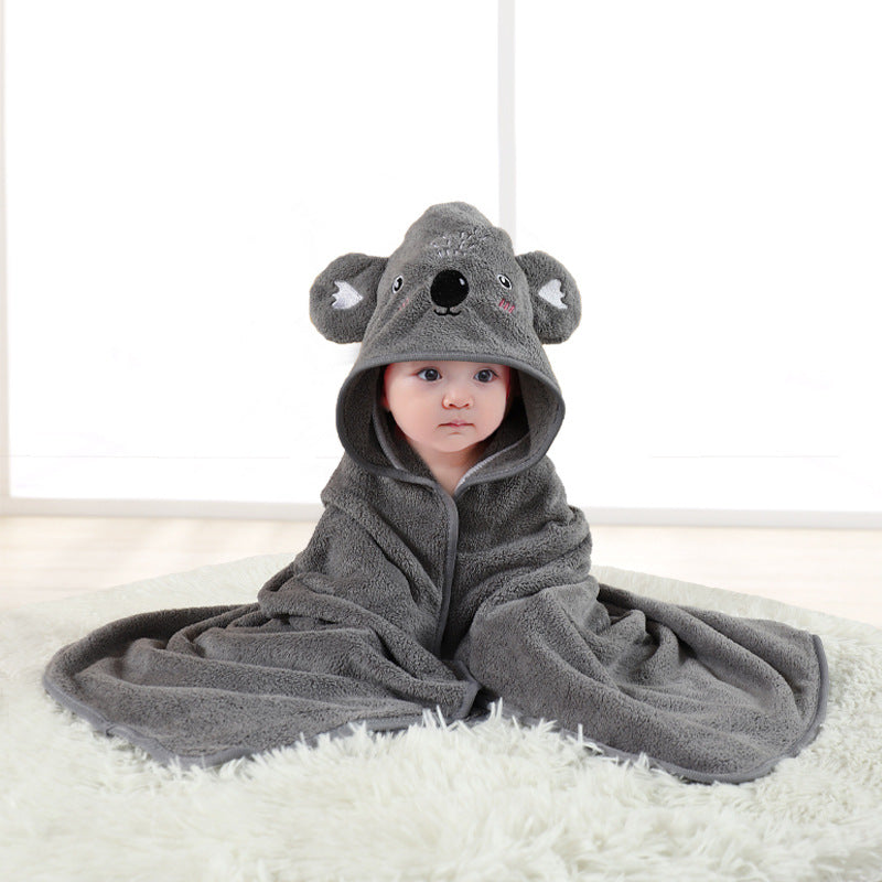 A baby wrapped in a cozy, gray koala-themed blanket with a hood featuring koala ears and face, sits on a soft white rug. The room is softly lit with a minimal background.