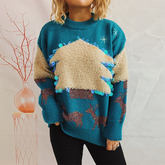 Person wearing a teal sweater with a large beige textured Christmas tree and sequins. The sweater also features reindeer patterns. Background shows a vase with branches.